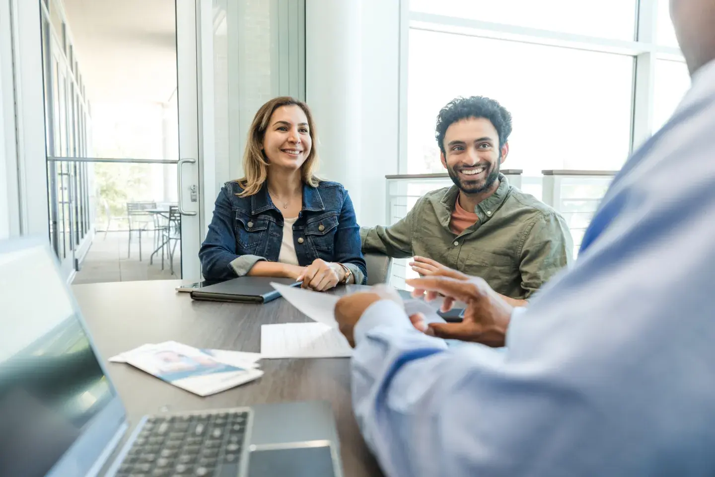 Smiling couple shaking hands with a professional across a desk in a bright office setting.